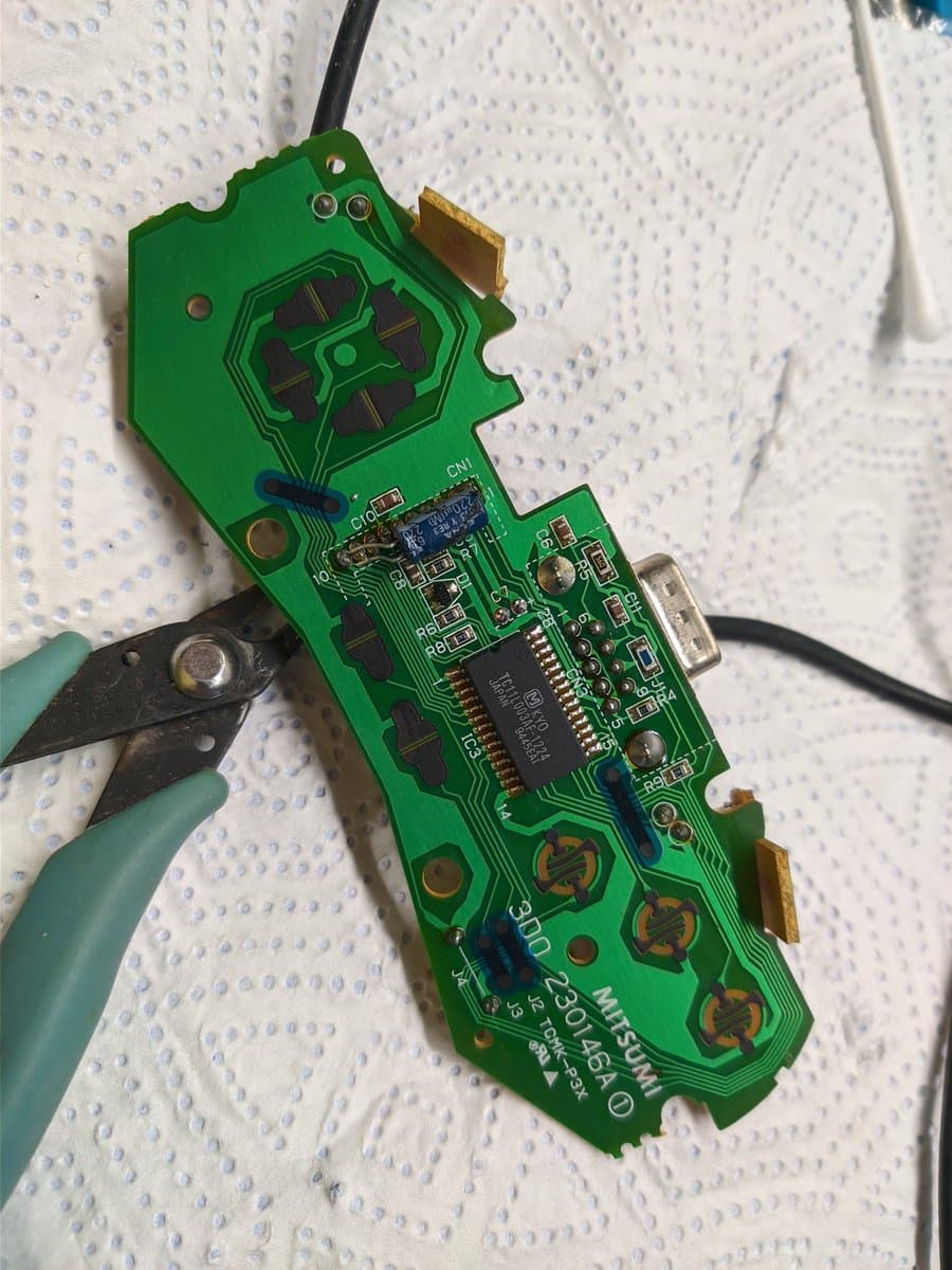 3DO controller PCB being recapped with tweezers, showing exposed capacitors and IC chips during refurbishment work.