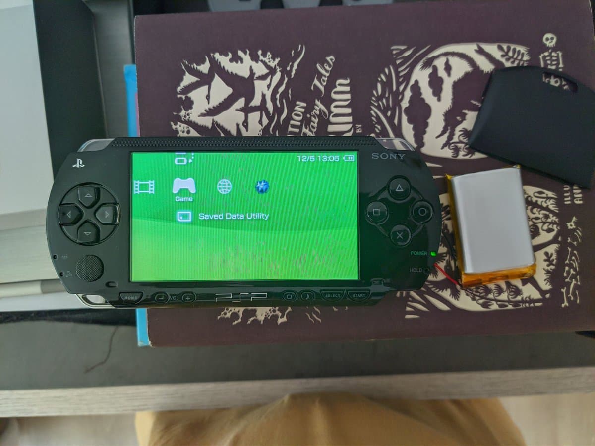 PSP-3000 displaying home menu with replaced battery beside device; battery cell visible with yellow protective tape during re-celling repair process.