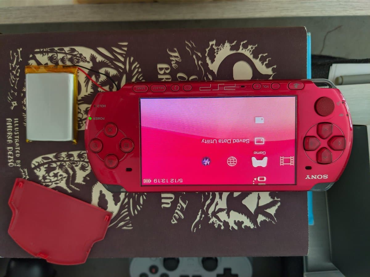 Sony PSP-3000 displaying home screen next to brown instruction manual and red battery cover during re-celling repair process.