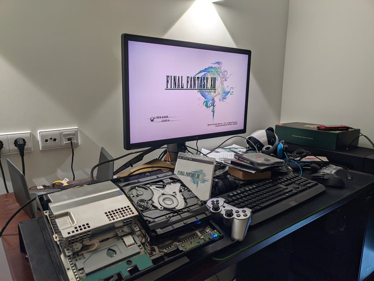 Disassembled PS3 CECHB00 motherboard and disc drive mechanism on black desk with controller and Final Fantasy XIII on monitor.