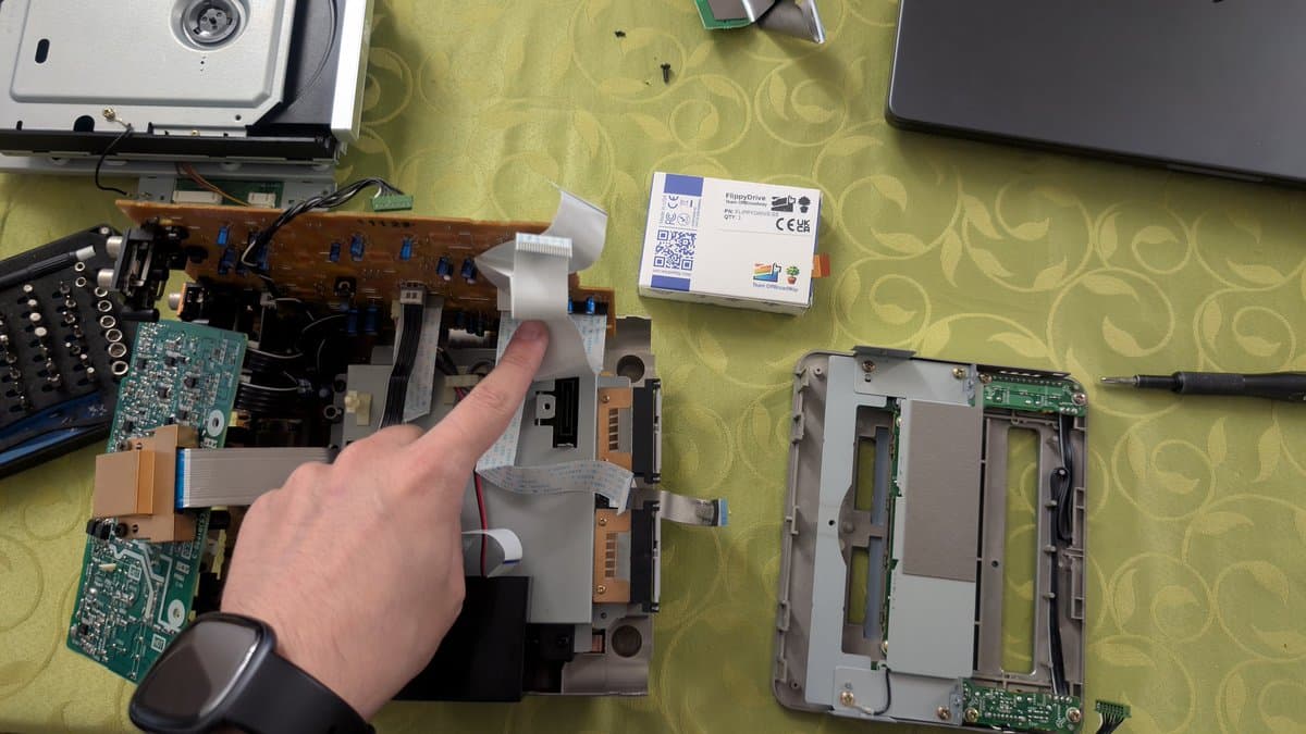 Technician's hand pointing to FlippyDrive flex cable in disassembled Panasonic Q optical drive during installation, showing opposite orientation versus standard GameCube.