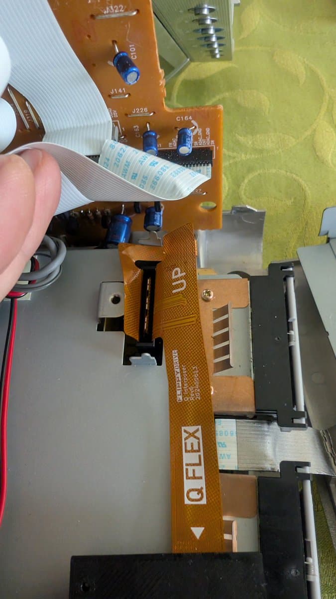 Hands holding flex cable above Panasonic Q optical drive board, showing reversed orientation compared to standard GameCube during FlippyDrive installation.