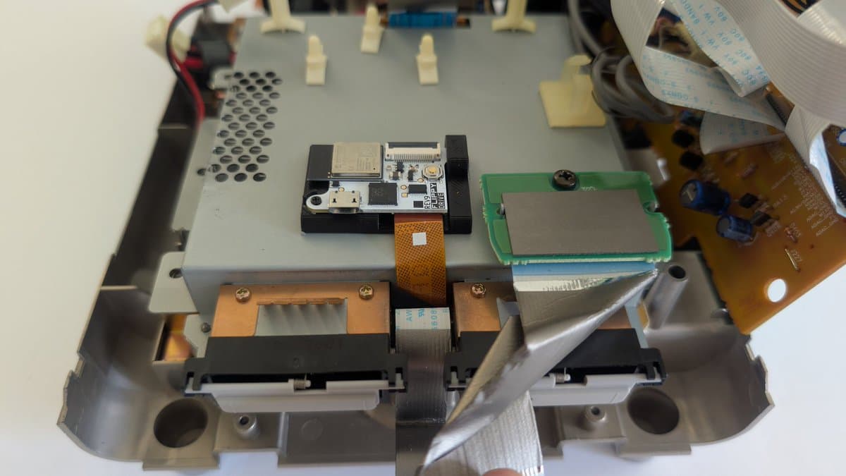 Panasonic Q internals showing FlippyDrive installation with flex cable routed above shielding, green PCB visible, orange copper heatsinks, black drive mechanism.