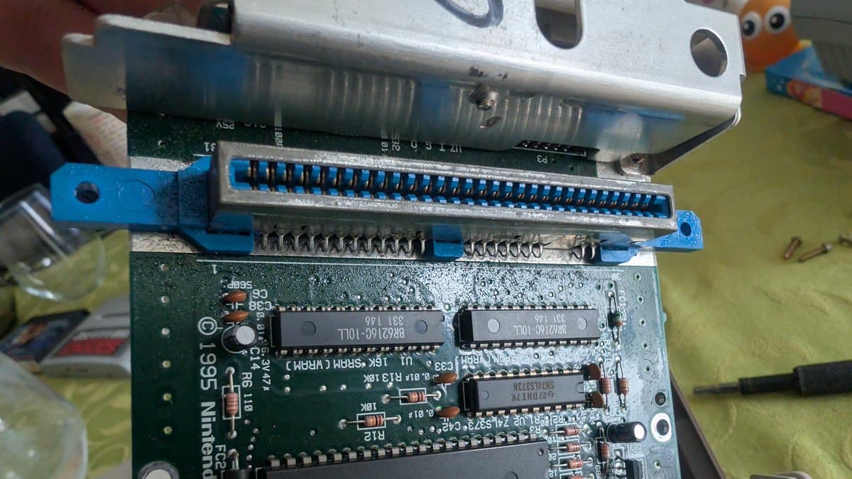AV Famicom motherboard with blue ribbon cable connector and visible IC chips during recap maintenance, dust and oxidation visible.