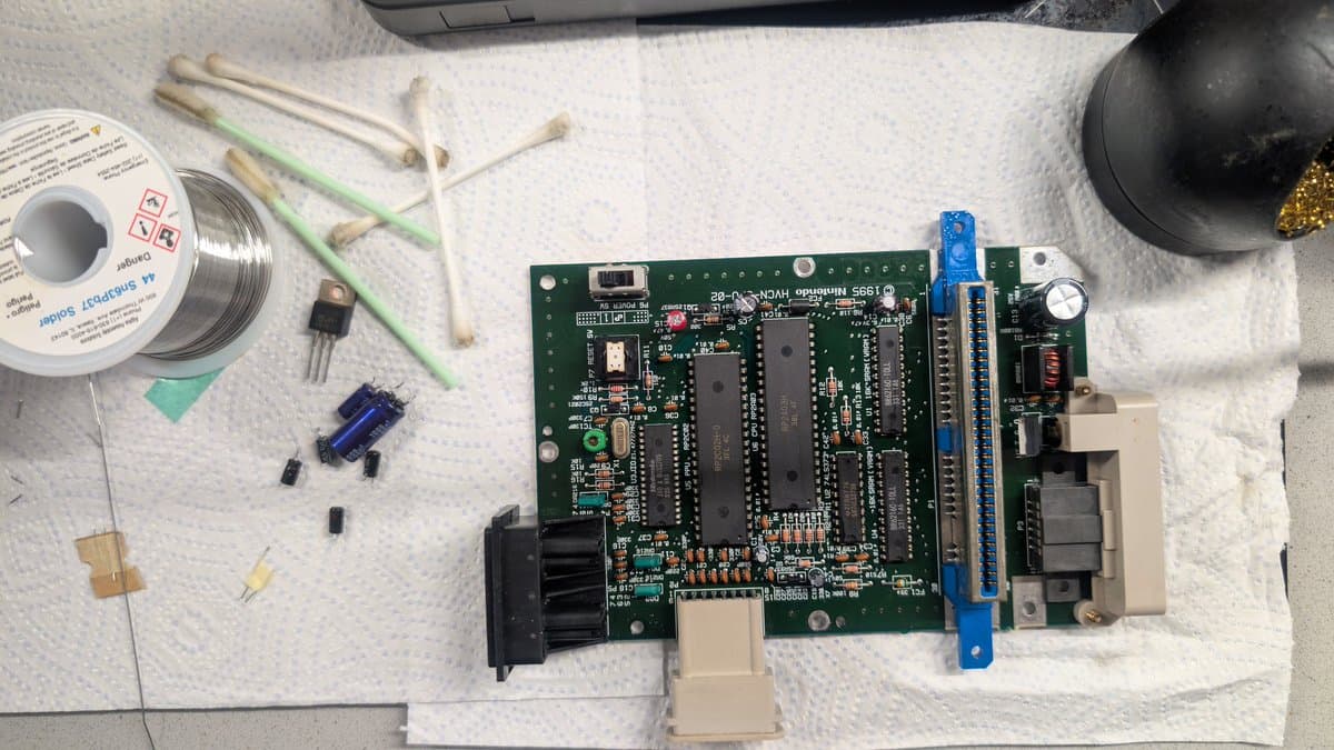 AV Famicom motherboard during recap repair, showing replaced capacitors, soldering iron, solder spool, and assorted electronic components on work surface.