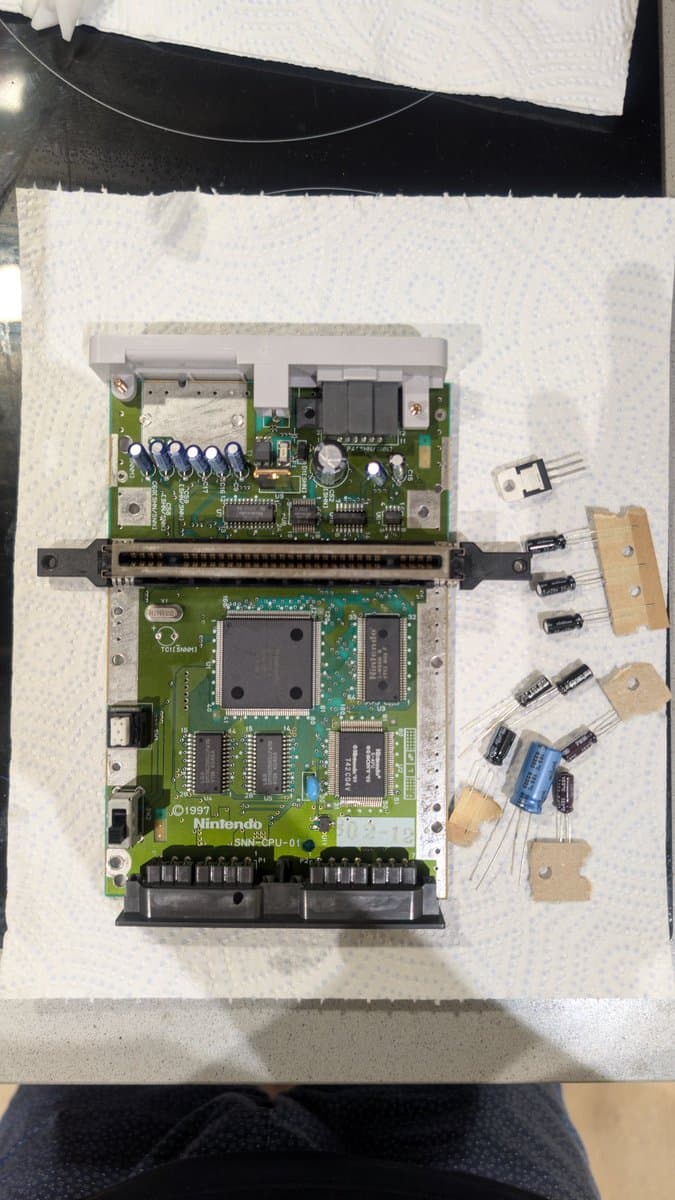 Disassembled Super Famicom Jr. mainboard with removed capacitors on wooden mounts, showing CPU board and power board separated.