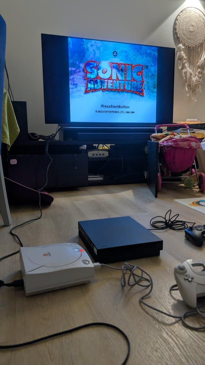 Sega Dreamcast VA0 startup screen showing Sonic Adventure burn-in test, with white Dreamcast and black console visible on wooden floor.