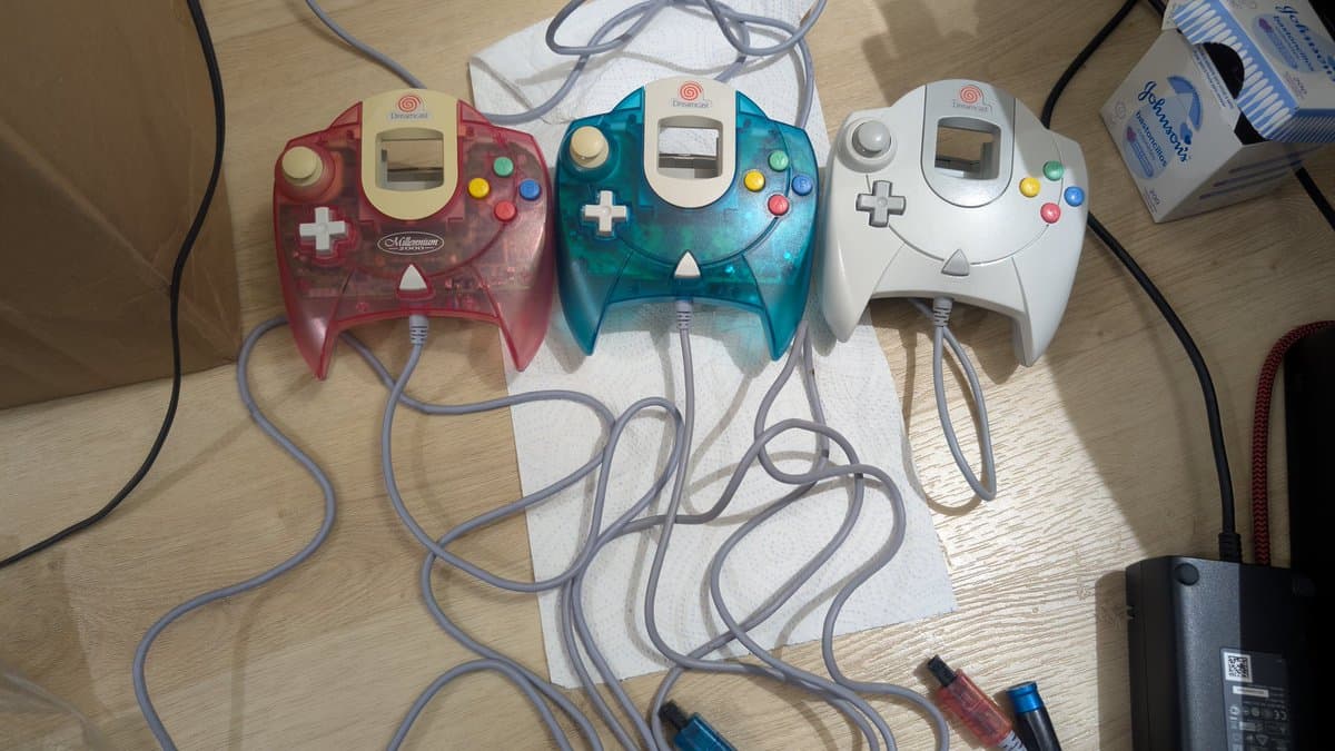 Three cleaned Dreamcast controllers in red, teal, and white arranged on a workbench with coiled cables.