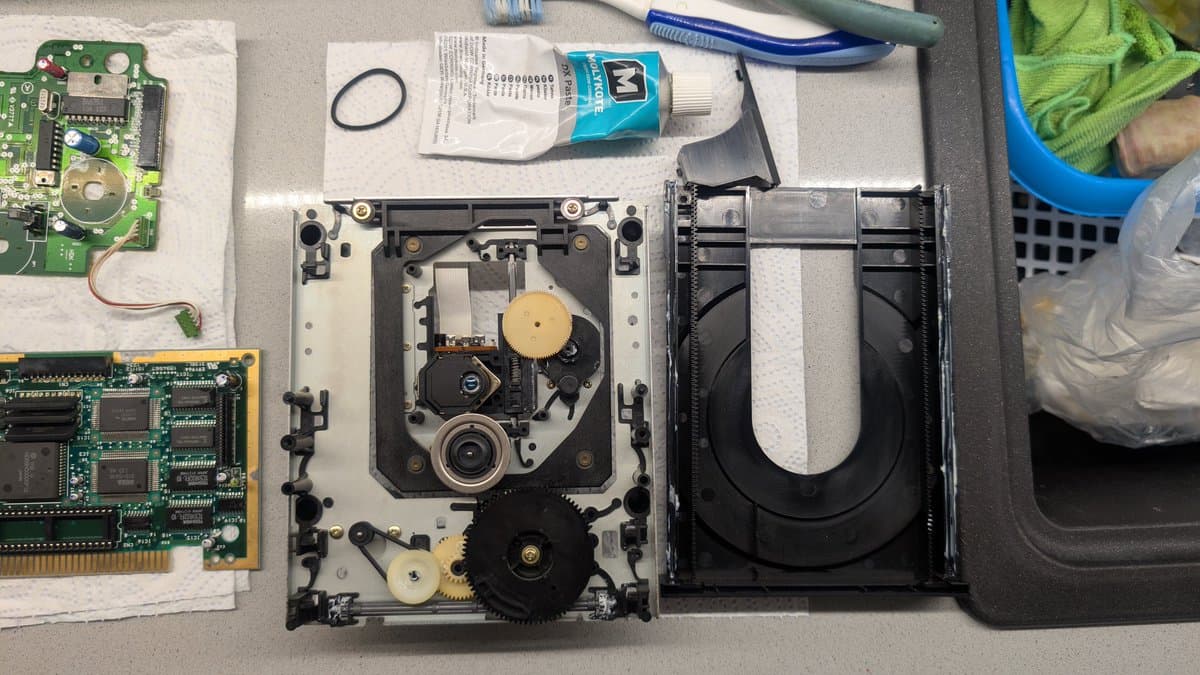 Disassembled Mega-CD 1 drive showing cleaned circuit boards, optical pickup mechanism, gears, and lubrication supplies on work surface.