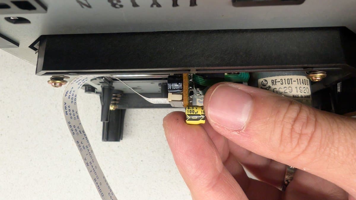 Technician's finger pointing at small yellow capacitor labeled "100μ" on Mega-CD circuit board; ribbon cable and green PCB visible nearby.