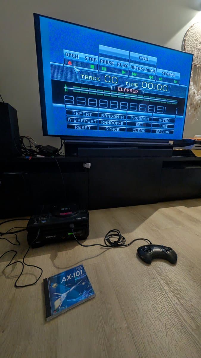Sega Mega-CD 1 displaying CD player menu on TV; console and controller visible; AX-101 game case on desk.