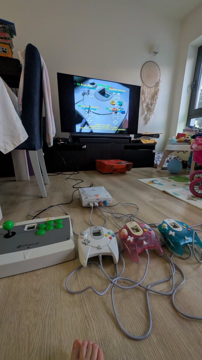 Dreamcast connected to TV running 240p test suite with four controllers plugged in; arcade stick and controllers on wooden floor during testing phase.