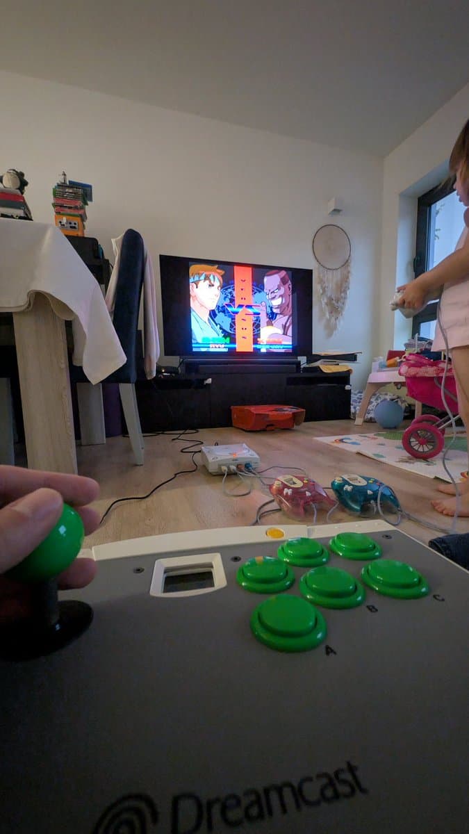 Dreamcast controller with green buttons being tested during final verification; Street Fighter Alpha 3 displayed on TV in background.