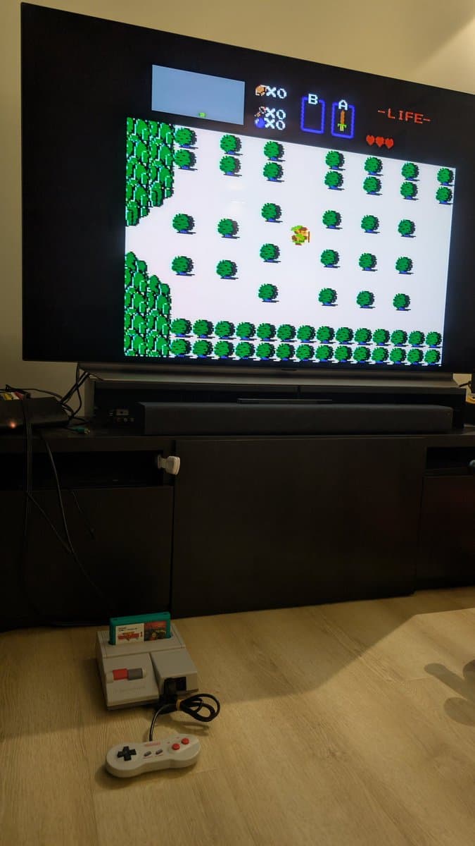 NES console with original controller displaying Legend of Zelda gameplay; game cartridges visible on floor below TV screen.