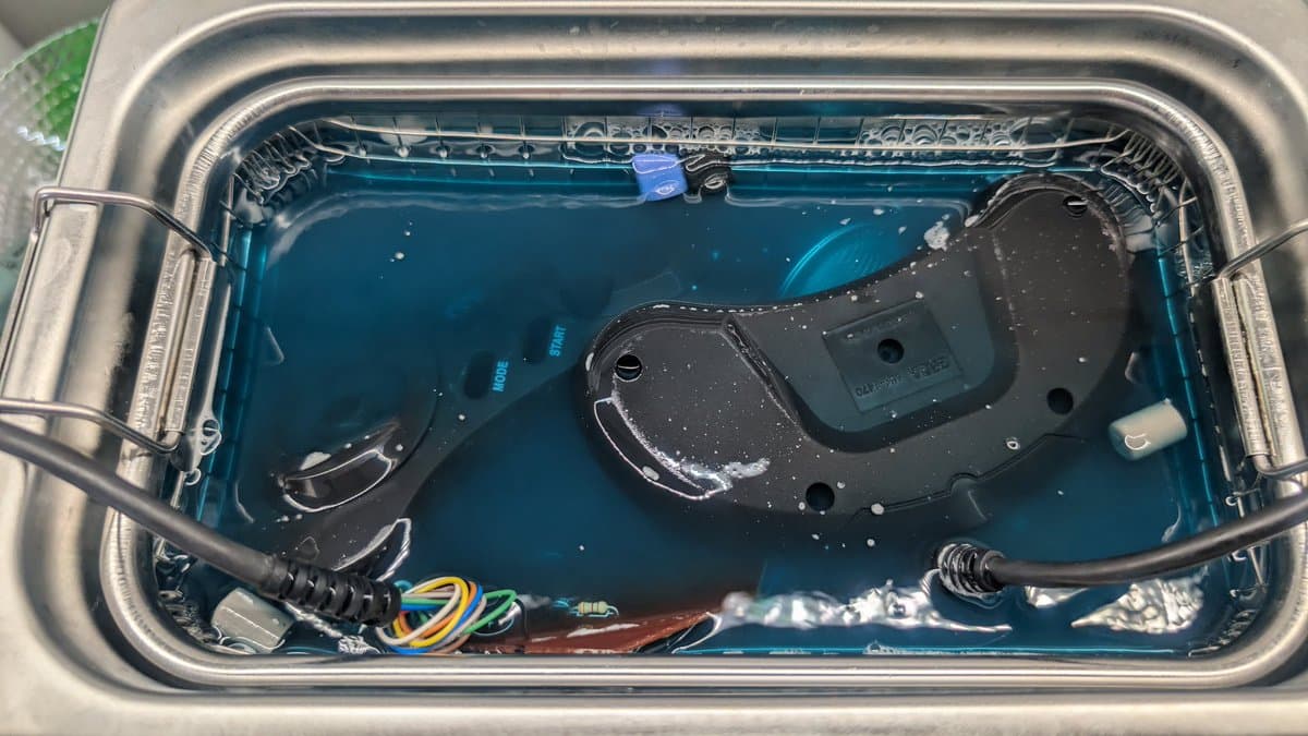Disassembled Mega Drive controller in ultrasonic cleaning bath with blue solution, showing internal circuit board and components during deep clean.