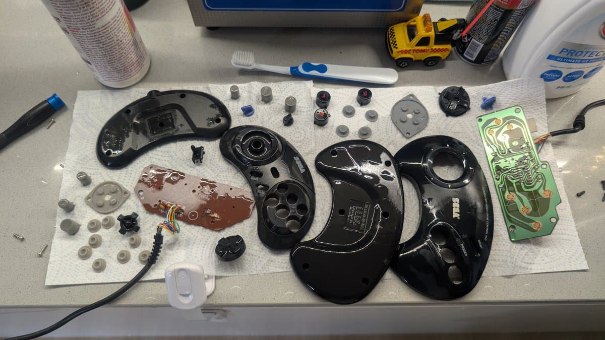 Disassembled Mega Drive controllers on white mat showing black plastic shells, circuit boards, button mechanisms, springs, and small components during deep cleaning restoration.