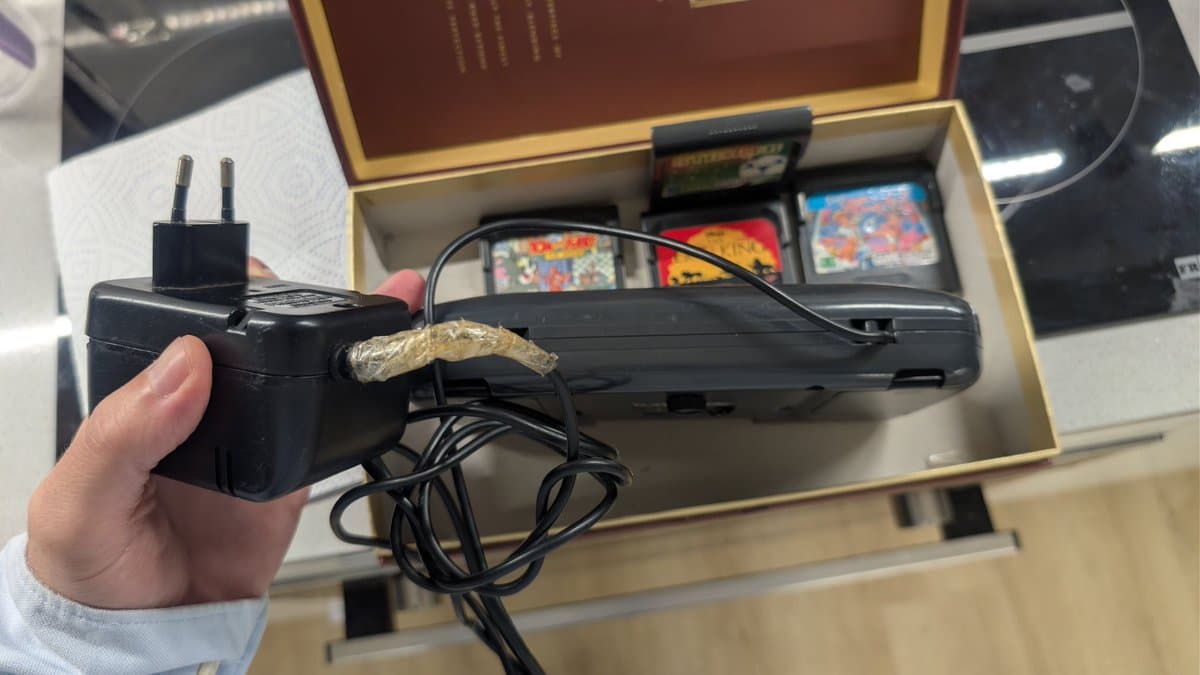 Hand holding black Game Gear AC adapter with attached transformer; gold foil repair visible on connector; console box with game cartridges in background.