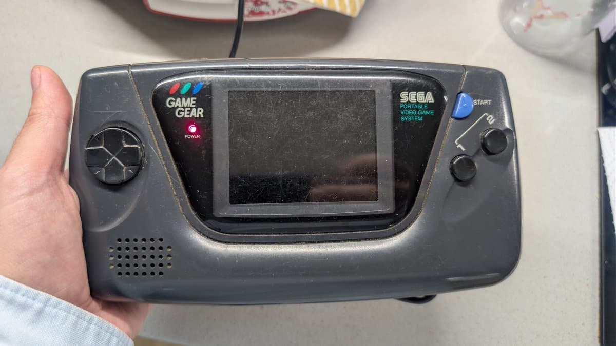 Hand holding a dark gray Sega Game Gear portable console with blank screen, showing power LED and controller inputs.