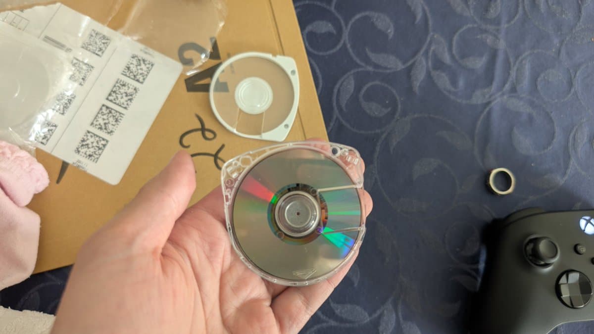 Hand holding cracked PSP disc with visible damage; disc case and QR code labels visible on cardboard surface.