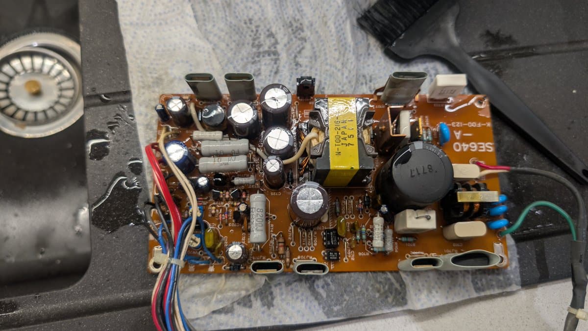 Opened Sharp X68000 PSU board showing brown PCB with multiple electrolytic capacitors, yellow transformer, and visible electrolyte residue from failed caps.