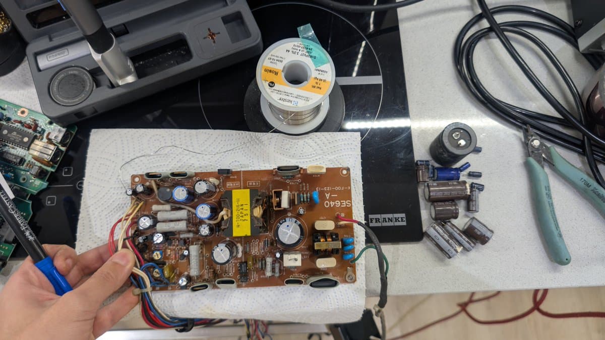 Sharp X68000 PSU board held in hand showing bulging capacitors, electrolyte damage, replaced caps, and repair tools on work surface.