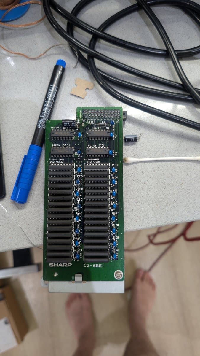 Sharp CZ-6BE1 RAM expansion board after recap, showing dual RAM chip arrays with blue capacitors, alongside marker and black cables on work surface.