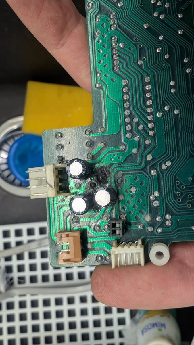 Close-up of damaged PCB section showing three electrolytic capacitors, burnt component, and corroded solder joints from failed capacitor on Sharp X68000.