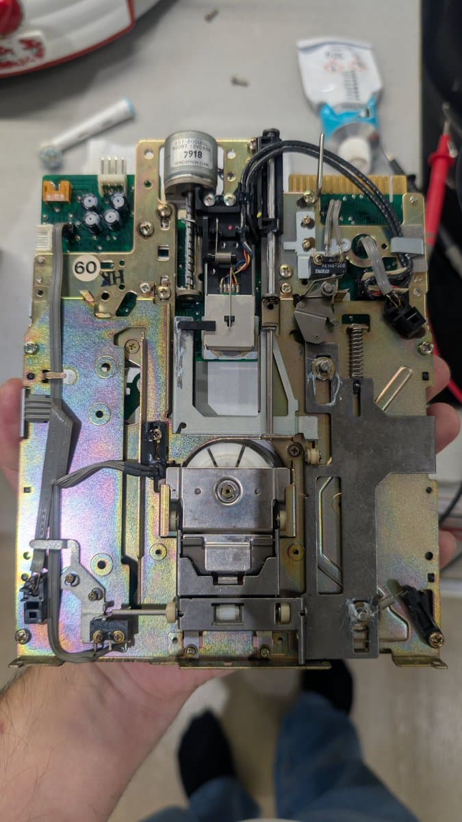 Sharp X68000 motherboard held in hand showing brass chassis, capacitors, transformer, motor assembly, and green PCBs during restoration work.