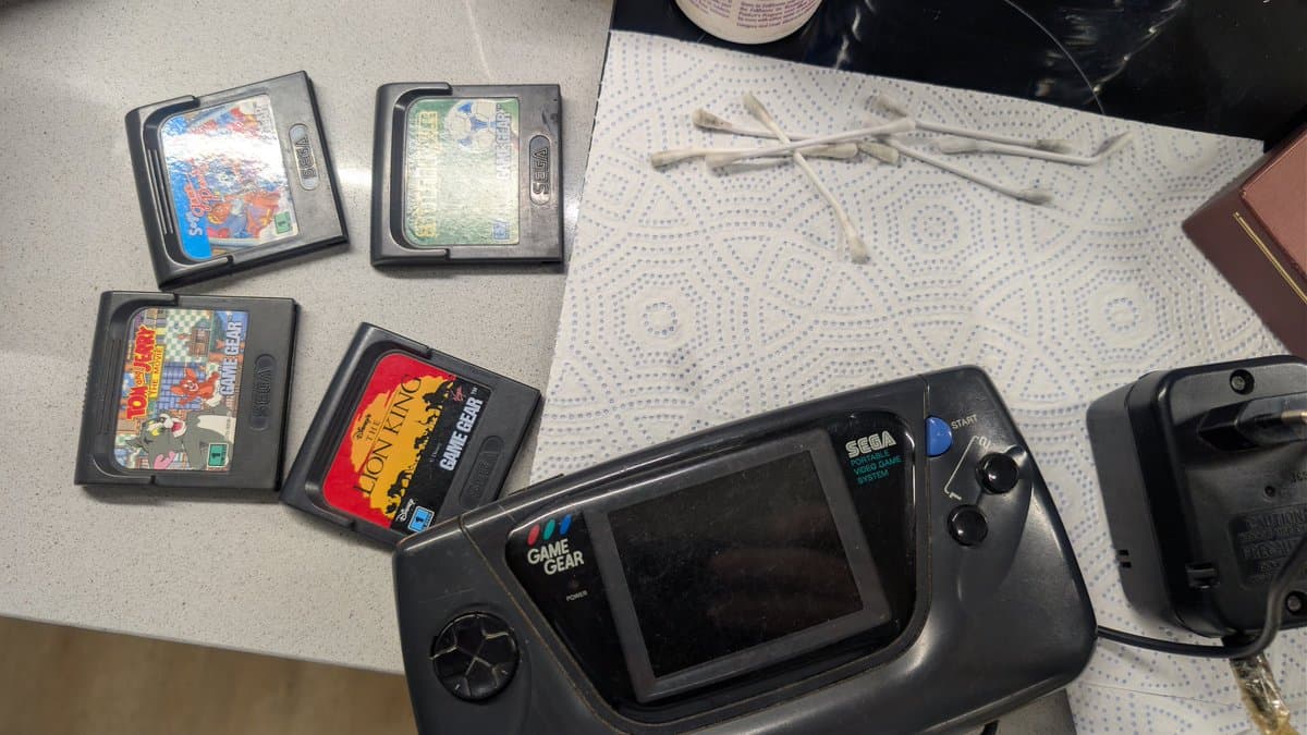 Sega Game Gear with four game cartridges arranged above it on a workbench with white dotted cloth, ready for cleaning and repair diagnostics.