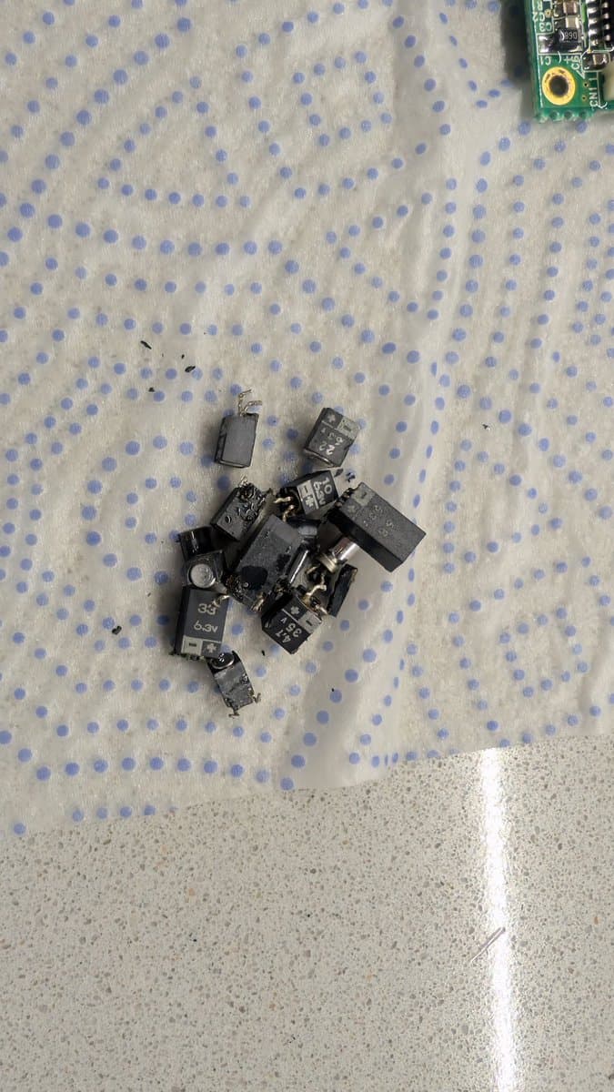 Pile of desoldered electrolytic capacitors from Sega Game Gear main board on dotted fabric, with circuit board visible top-right.