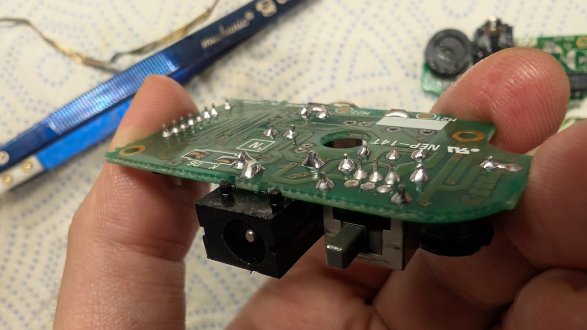 Close-up of Game Gear power board being held, showing reflowed solder joints on green PCB with black power connector and soldering iron in background.