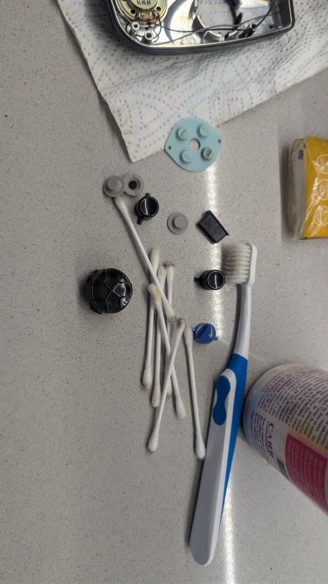 Disassembled Game Gear buttons and components laid out: rubber pad, contacts, springs, blue toothbrush, and cleaning supplies.