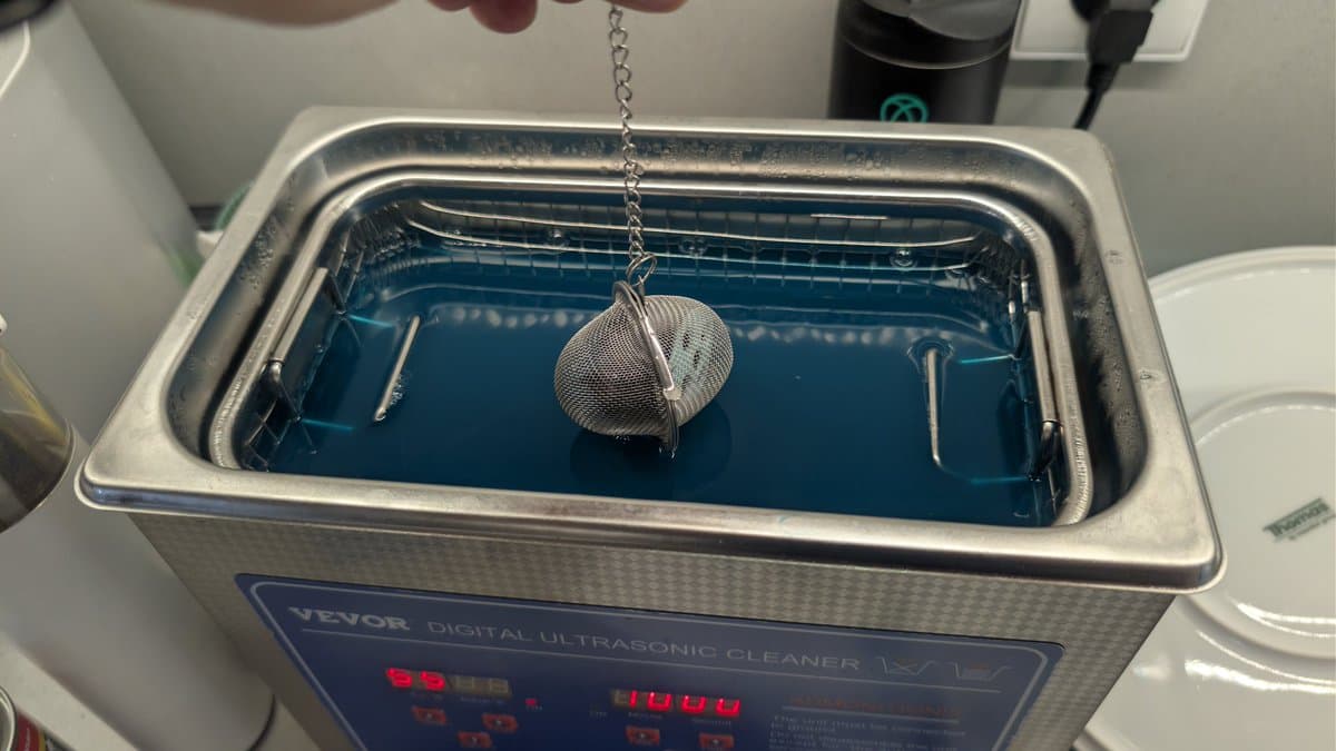 Mesh tea infuser ball suspended in blue liquid inside JEVOR ultrasonic cleaner, cleaning Game Gear buttons.