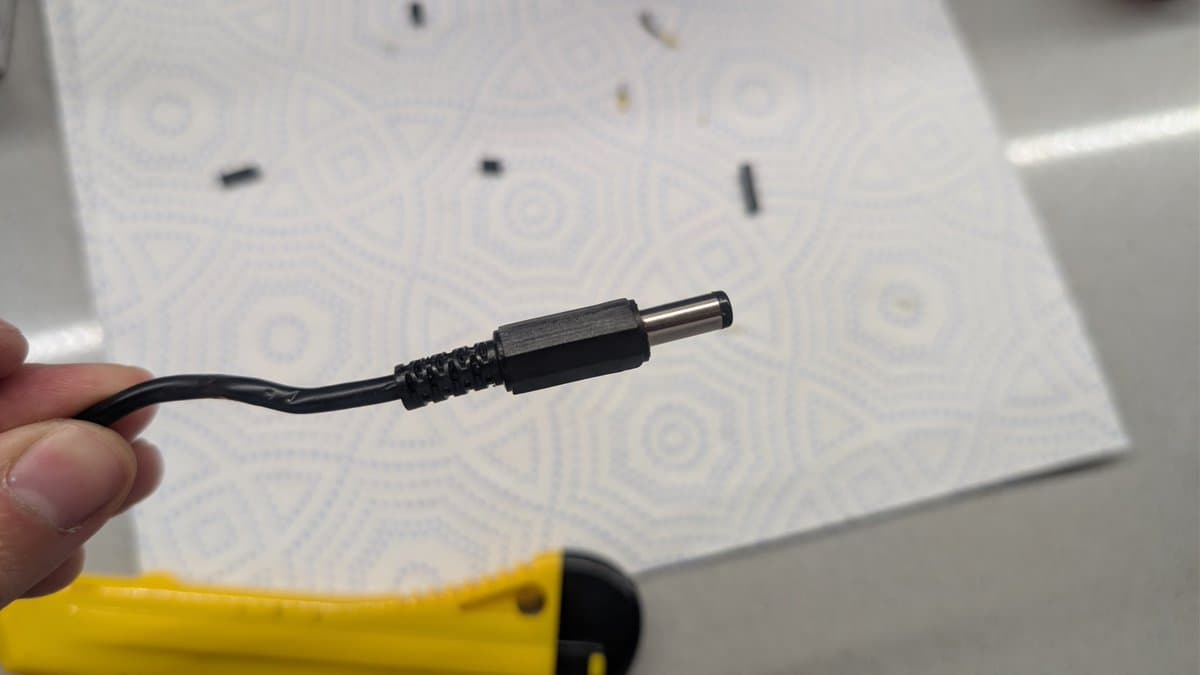 Close-up of barrel connector and black power cable for Sega Game Gear external PSU restoration against patterned white background.