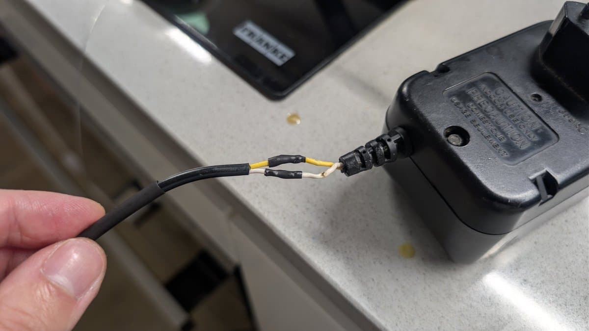Close-up of Game Gear power cable repair: exposed wires with yellow and black heat shrink tubing partially applied, connector visible, held over work surface.