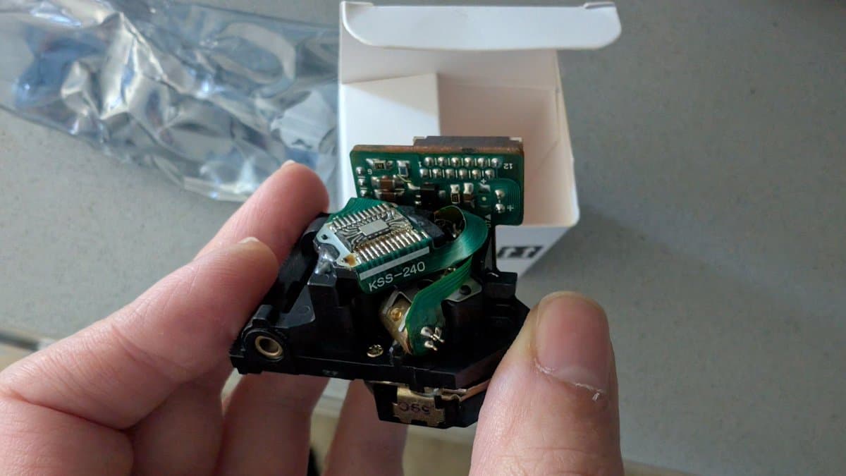 Hand holding KSS-240A optical pickup with green circuit board, black housing, and metal components over white box containing additional parts.