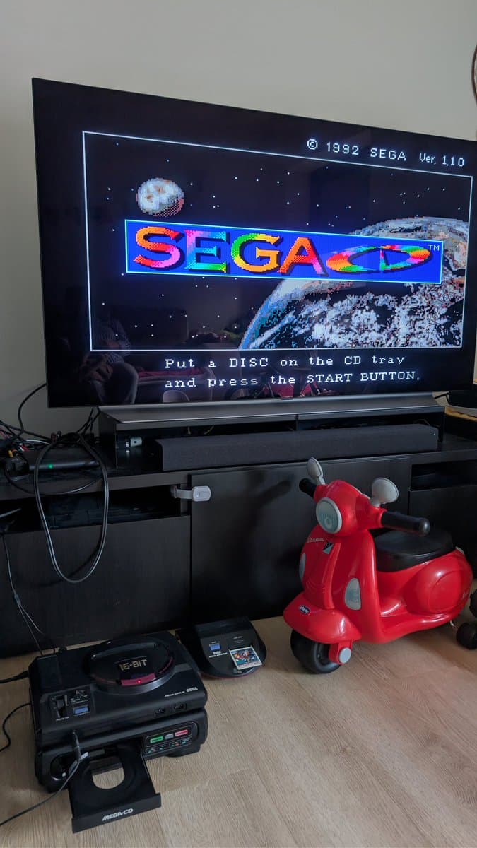 Sega Mega-CD 1 with disc tray open displaying SEGA boot screen, accompanied by original optical pickup component on wooden desk.