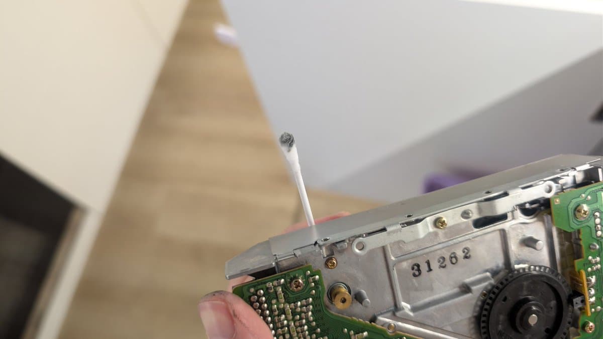 Fujitsu FM Towns Marty floppy drive PCB being cleaned during restoration; tweezers removing debris near motor shaft and green circuit board components.