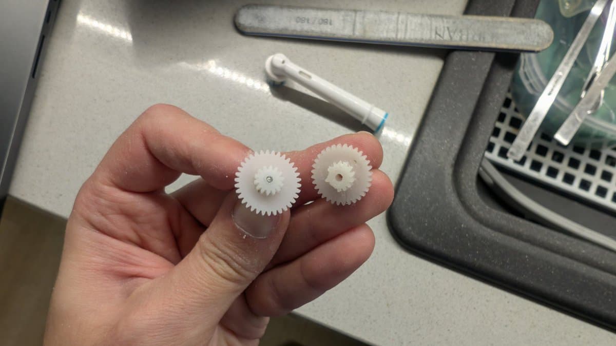 Two white plastic gears held in hand; left gear shows filed teeth where two smaller gears were cut and glued together to replace missing teeth on original gear.