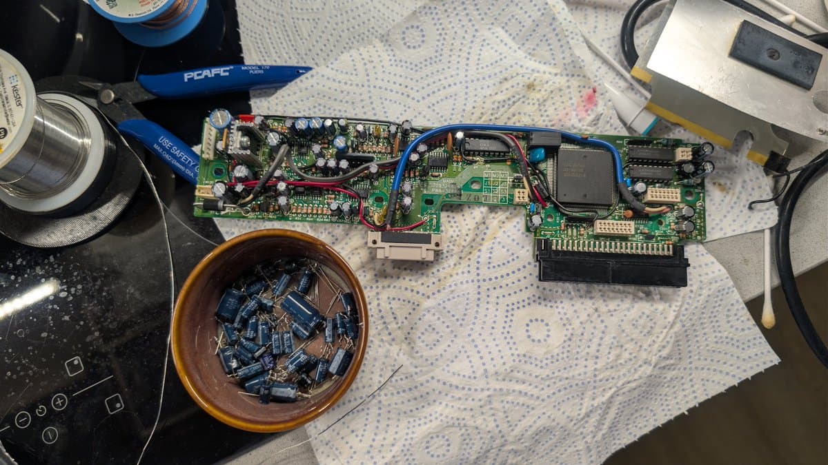 PC Engine IFU-30 main board with removed blue capacitors in bowl, soldering iron, and multimeter during recap repair.