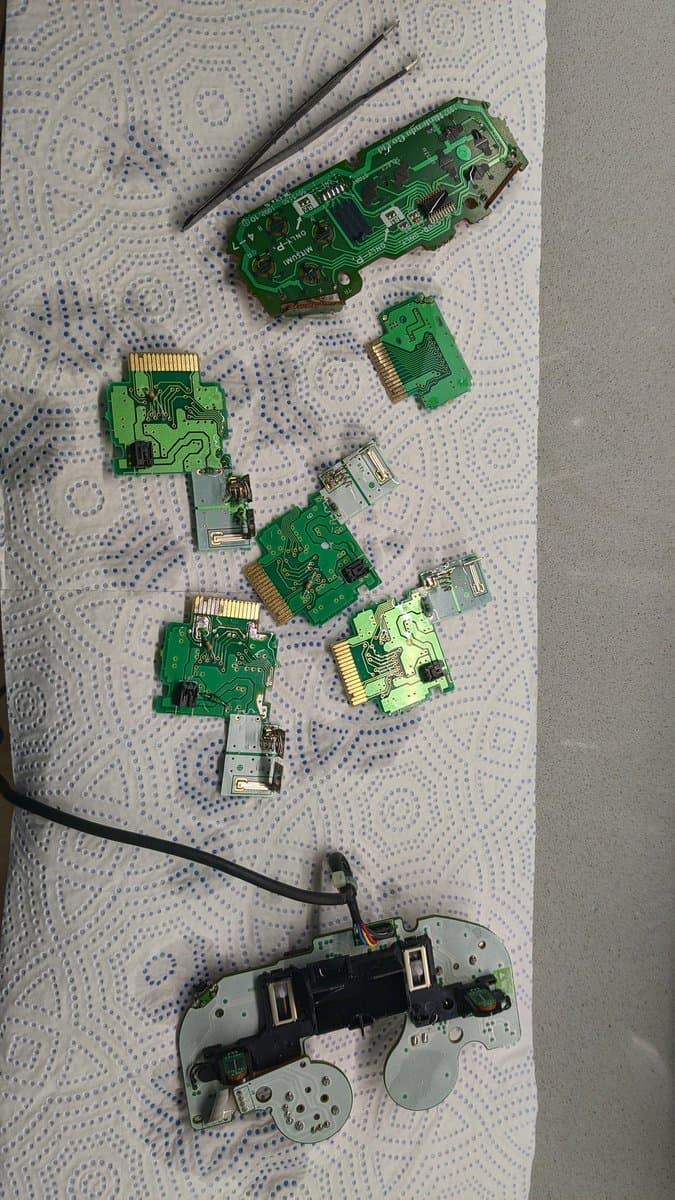 Disassembled controller PCBs and rumble pack boards laid out on patterned fabric, cleaned via ultrasonic cleaner with tweezers nearby.
