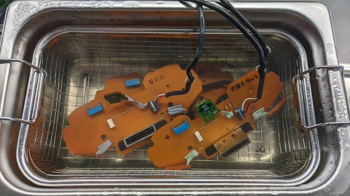 Orange PCBs and controller shells in ultrasonic cleaner basket with soapy water, showing capacitors, connectors, and circuit traces after cleaning.