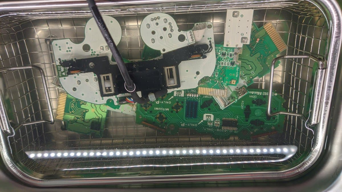 Nintendo controller PCB and plastic components in ultrasonic cleaner basket, showing white shells, green circuit boards, and LED strip mid-cleaning.
