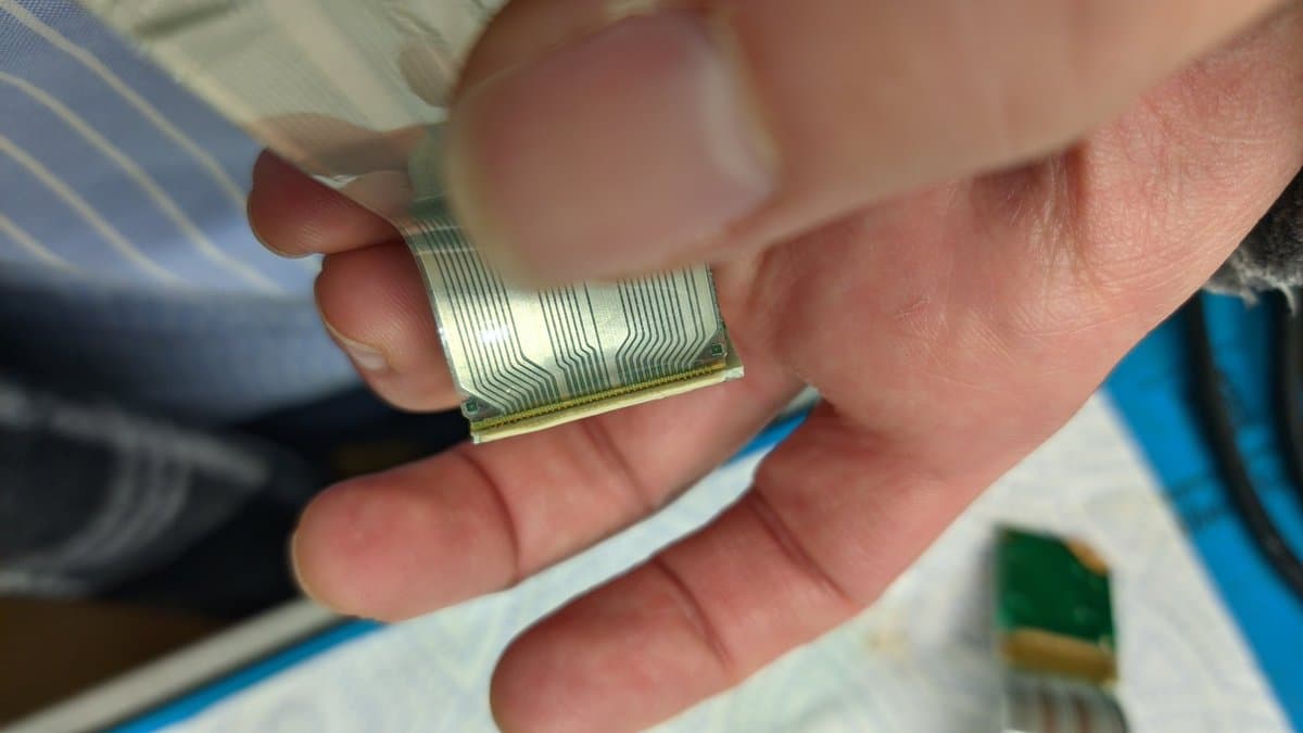 Hands holding a flexible ribbon cable with gold connector contacts and fine parallel traces, separated from Virtual Boy LED array PCB during refurbishment.