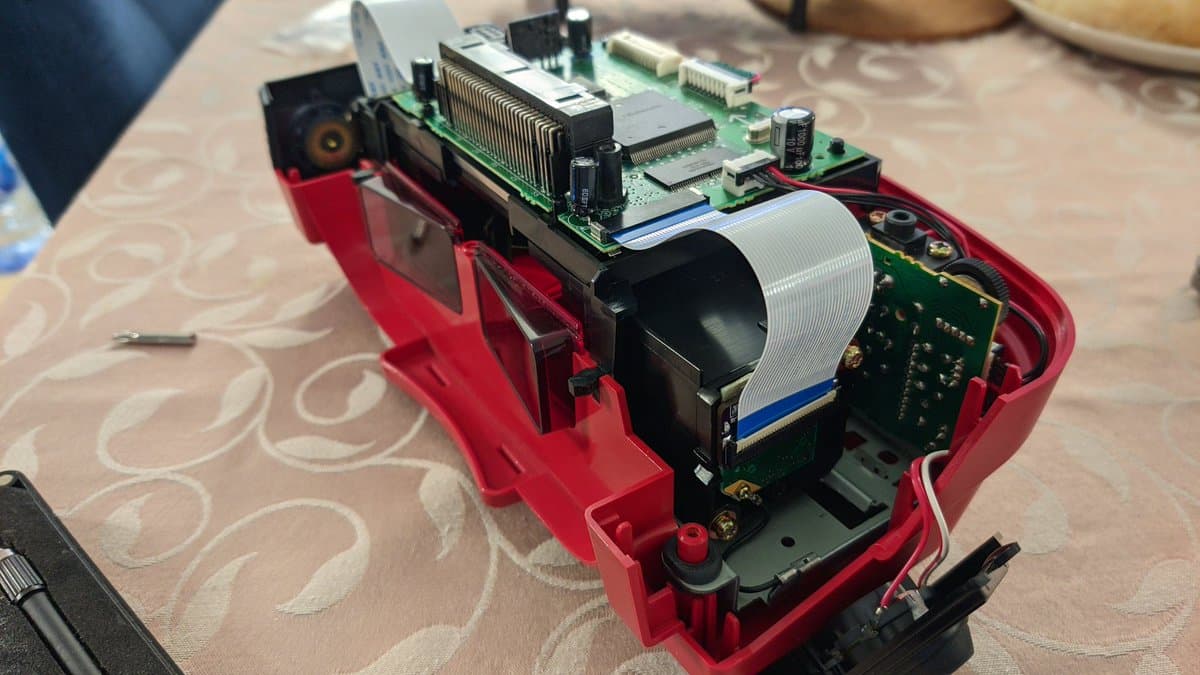 Virtual Boy motherboard with ribbon cable mounted in red jig, showing uneven pressure from single-screw mounting design during VB 4.0 installation.