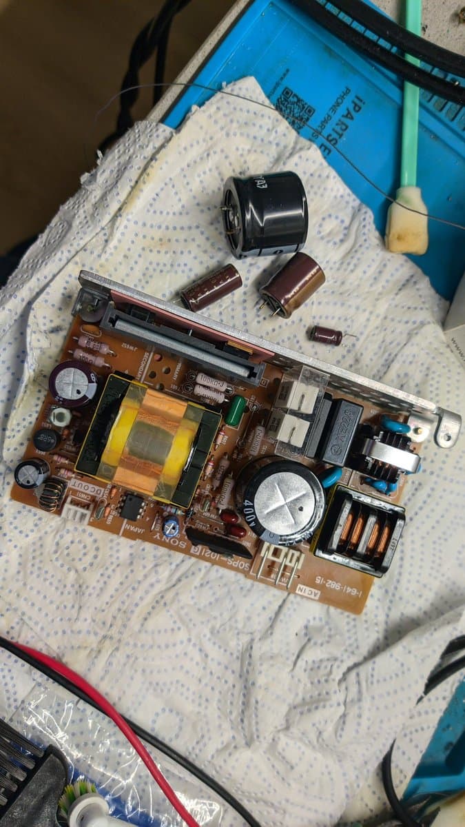 Sony PVM-9041Q PSU board with removed capacitors and trimmer potentiometers on work surface, awaiting recapping and component replacement.