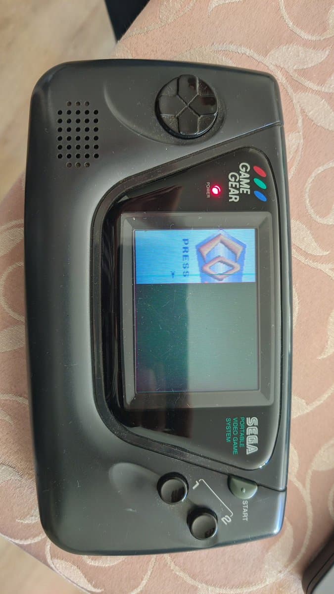 Sega Game Gear with IPS display mod showing Sega logo boot screen, speaker grille visible top left, glass lens installed, START button visible bottom right.