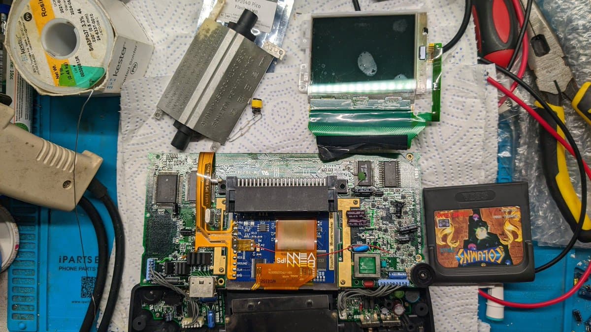 Game Gear motherboard with IPS screen, FM module, and speaker installed; torn flex cables visible, clock line bodge wire repair in progress.