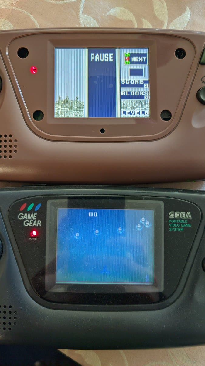 Two Sega Game Gear handhelds stacked vertically; top unit shows IPS screen displaying pause menu with score/block/level; bottom unit shows stock screen with blue display.