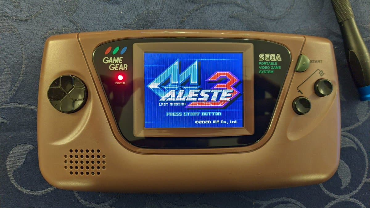 Gold Sega Game Gear with IPS display mod showing Adeste Last Mission boot screen, glass lens installed, speaker grille visible, d-pad and buttons on sides.